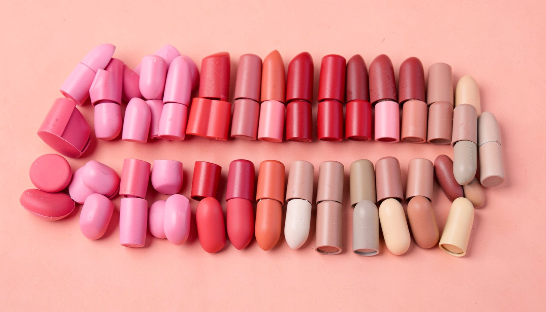 Rows of lipstick bullets in pink, red, and nude tones arranged on a blush-coloured background