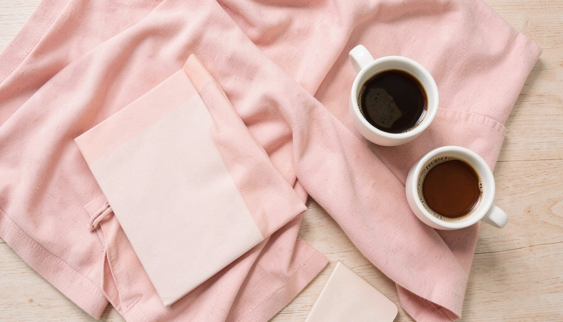 Soft pastel fashion flat-lay with blush-toned fabrics, a notebook, and a coffee cup on a light wood surface, warm and inviting mood