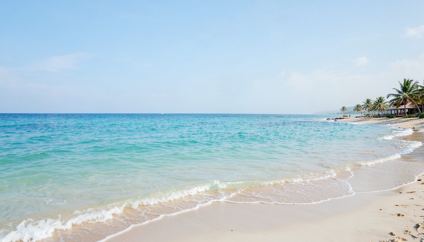 Wide-angle view of a calm turquoise ocean meeting a sandy shoreline under a soft blue sky, with gentle waves and a peaceful resort atmosphere